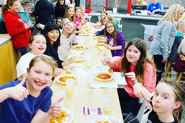 Group of children enjoying a meal at a long table in a kitchen setting.