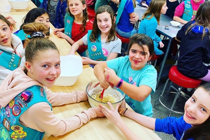 Group of girls wearing badges, mixing ingredients in a kitchen class, smiling at the camera.