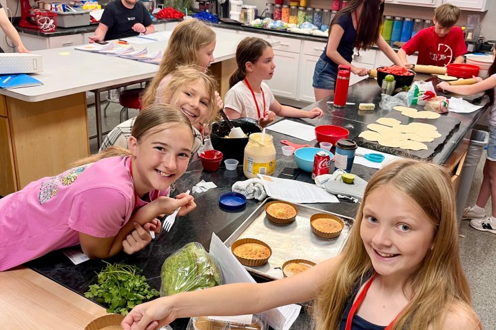 Children cooking and smiling in a bright kitchen with ingredients on the counter.