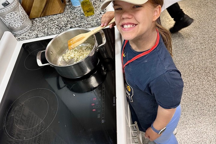 Person in cap smiling, stirring onions in a pot on a stove, surrounded by kitchen items.