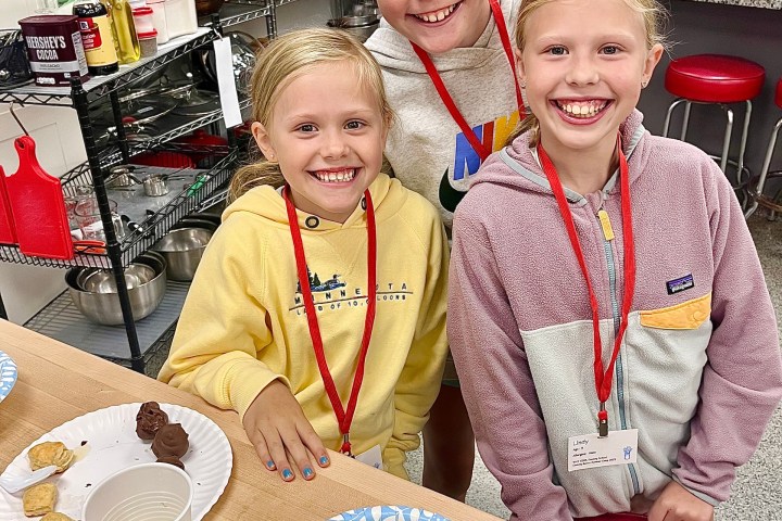 Three smiling girls with red lanyards at a kitchen counter with food and drinks.