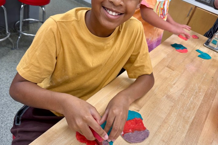 Smiling child cutting colorful dough at a wooden table with another child in the background.