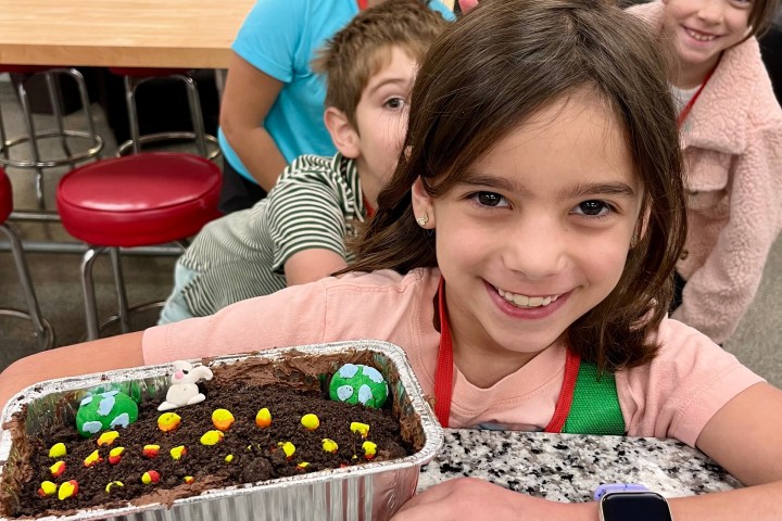 Smiling kids in a kitchen holding a decorated cake in a foil tray.
