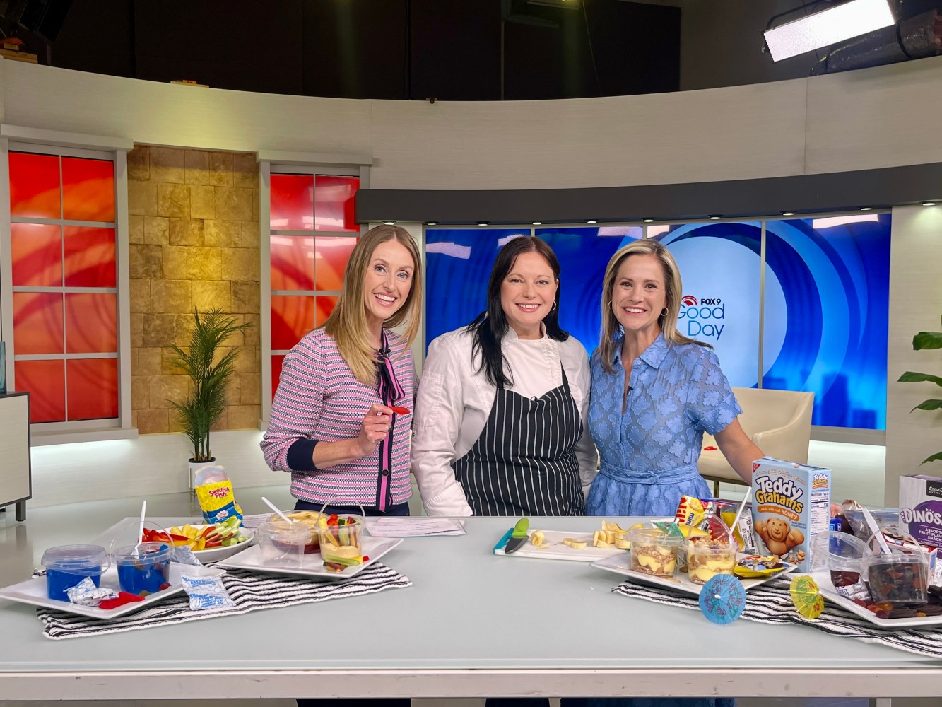 Three women posing in a TV studio kitchen with dessert trays.